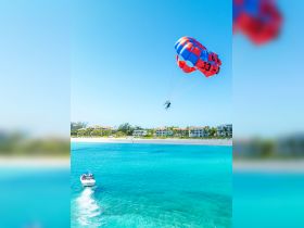 A thrilling parasailing experience over stunning turquoise waters, with a boat below and beautiful beachside resorts in the background — perfect for adventure seekers enjoying water sports.