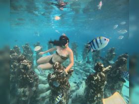 A snorkeler explores an underwater art installation surrounded by vibrant fish, showcasing the beauty of marine life and underwater adventures.
