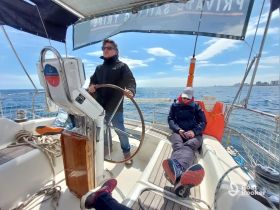 A sailor navigates the helm while a guest relaxes in the cabin of a sailboat, showcasing the comfortable interior space perfect for leisurely sailing.