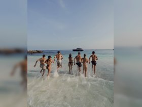 A lively group of friends runs into the water from the beach, with a luxurious yacht anchored nearby — capturing the essence of summer fun and adventure on the water.