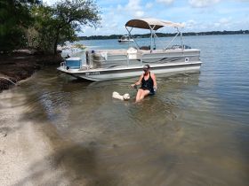 A woman enjoys a relaxing day at the beach with her dog, wading in the water next to a pontoon boat — a perfect scene for pet-friendly outings on the water.