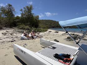 A couple relaxes on the beach next to a catamaran, enjoying the sun and scenery, with snorkeling gear ready for underwater exploration.