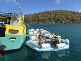 A lively group enjoys drinks on a floating mat anchored near a colorful boat, perfect for a fun day on the water.