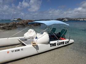A catamaran named 'The Cove Cat' is docked on a sandy beach, surrounded by clear waters and distant sailboats, perfect for a relaxing day on the water.