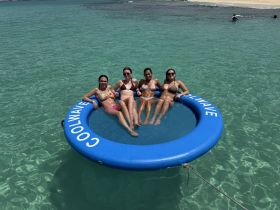 A group of friends enjoys a sunny day on a floating mat in crystal-clear waters, perfect for relaxation and fun at the beach.