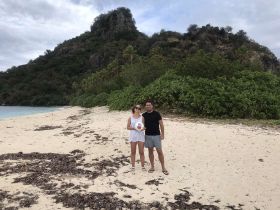 A couple poses on a secluded beach, surrounded by lush greenery and rocky hills — a perfect snapshot of their island getaway.