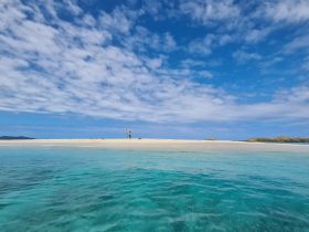 A person stands on a pristine beach, arms raised in celebration against a backdrop of clear blue skies and turquoise waters — a perfect moment captured during a tropical getaway.