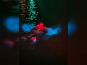 An unclear image of a person swimming in dark waters, illuminated by colorful lights. The low visibility makes it difficult to discern details.