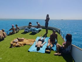 A lively group enjoys a sunny day on the deck of a boat, relaxing and sunbathing on the turquoise waters — perfect for a fun day out at sea.