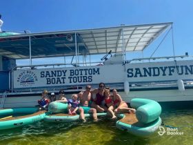 A joyful family enjoys a sunny day on the water with Sandy Bottom Boat Tours, featuring swimming and fun activities on floating mats.