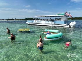 A lively group enjoys a sandbar stop, swimming and relaxing around the pontoon boat 'Sandyfun' — perfect for family fun in crystal-clear waters.