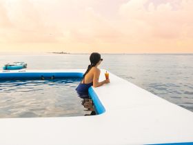 A woman enjoys a cocktail while relaxing in a floating pool at sunset, with a kayak in the background — a perfect scene for unwinding on the water.