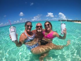 A joyful group enjoys a refreshing swim in crystal-clear waters, holding cocktails and celebrating under the sun — a perfect beach day experience.