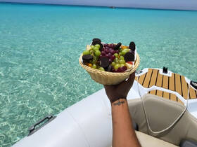 A hand holds a basket of fresh fruit and snacks against a stunning turquoise backdrop, perfect for enjoying on a boat trip or beach day.
