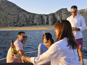 A lively group enjoys drinks on a boat at sunset, capturing the moment with their phones against a stunning coastal backdrop.
