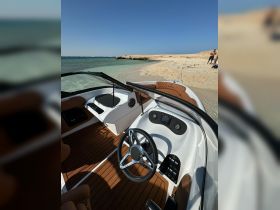 A well-designed helm area of a boat, showcasing the steering wheel and console, with a sandy beach and people in the background — perfect for a day of fun on the water.