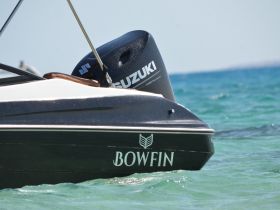 A close-up view of the back of a boat named 'BOWFIN' with a Suzuki outboard motor, showcasing its sleek design against the clear blue waters.