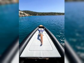 A person relaxes on the bow of a boat, soaking up the sun against a backdrop of clear blue waters and lush greenery — an ideal scene for a tranquil day on the water.
