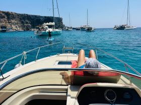 A relaxing day on the water, featuring a person lounging on a boat while others enjoy paddleboarding in a picturesque bay surrounded by cliffs.