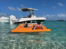 A couple relaxes on an orange floating mat in crystal-clear waters, with boats anchored nearby — a perfect scene for a fun day on the water.