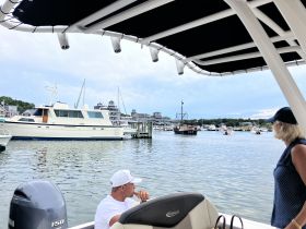 Guests enjoy a scenic view of the marina while relaxing on their boat, surrounded by various vessels and charming waterfront buildings.