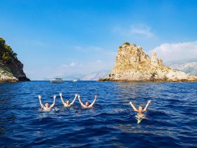 A group of friends joyfully swims in crystal-clear waters near a scenic island, with a boat in the background — a perfect snapshot of summer fun and adventure.