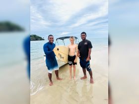 A young boy poses with two instructors on the beach, holding a surfboard, showcasing a fun surfing experience in a tropical setting.