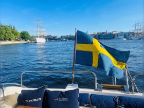 A picturesque view of a boat with the Swedish flag waving, set against a backdrop of a historic tall ship and the vibrant cityscape, perfect for showcasing maritime adventures in Sweden.