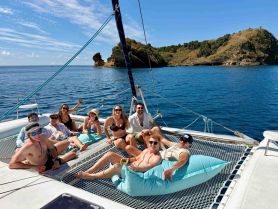 A lively group enjoys a sunny day aboard a catamaran, lounging on the netting with drinks in hand — perfect for a fun day on the water.