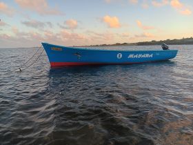 A vibrant blue fishing boat named 'MAYARA' anchored in calm waters, showcasing its colorful design against a serene backdrop of clouds and shoreline.