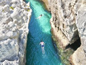 Two swimmers enjoy the crystal-clear waters of a scenic channel, surrounded by stunning rock formations — a perfect spot for a refreshing swim.
