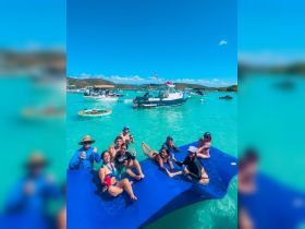 A lively group enjoys a fun day on the water, lounging on a floating mat with drinks in hand, surrounded by boats in a beautiful turquoise lagoon.