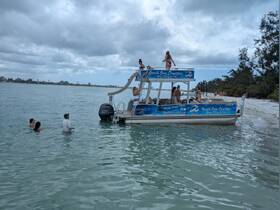 A lively group enjoys a beach day aboard a pontoon boat, with some swimming in the water and others relaxing on deck — perfect for a fun day out on the water.
