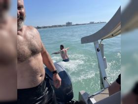 Two men enjoy a sunny day on the water, with one swimming and the other smiling on the boat — a perfect moment of fun and relaxation.