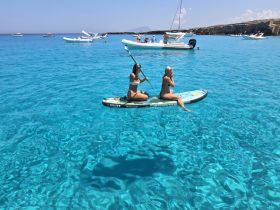 Two friends enjoy paddleboarding in crystal-clear turquoise waters, surrounded by boats — a perfect summer day on the water.