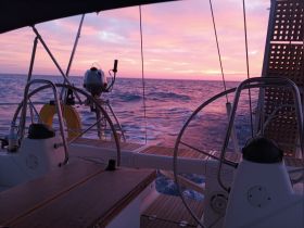 A serene view from the helm of a sailboat at sunset, showcasing the beautiful colors of the sky reflected on the water — perfect for a peaceful sailing experience.