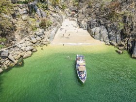 A scenic view of a boat anchored near a secluded beach, with people enjoying the sun and swimming in the clear waters — a perfect getaway for beach lovers.