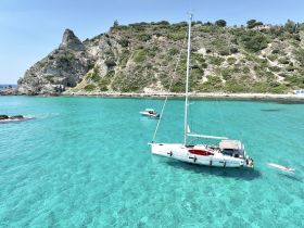 A beautiful sailing scene featuring a yacht anchored in crystal-clear turquoise waters, with guests enjoying paddleboarding and the stunning island backdrop.