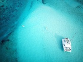 An aerial view captures a serene scene of a catamaran anchored in stunning turquoise waters, with a paddleboarder enjoying the calm surroundings — a perfect snapshot of a tropical getaway.