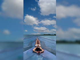 A woman relaxes while sunbathing on a transparent floating platform in serene waters, surrounded by beautiful blue skies and fluffy clouds — a perfect summer getaway.