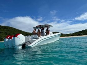 A group enjoys a sunny day on a sleek boat, surrounded by turquoise waters and a picturesque beach backdrop — perfect for a fun day on the water.