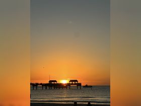 A stunning sunset view over a beach pier, with silhouettes of people enjoying the evening and a boat passing by — a perfect scene for relaxation and coastal adventures.