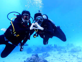 A couple shares a romantic moment underwater while scuba diving, forming a heart shape with their hands amidst vibrant marine life — a perfect snapshot of adventure and love.