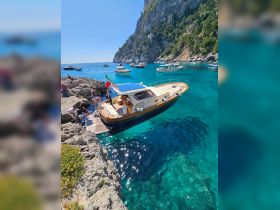 A lively scene at Capri with people interacting near a boat on a rocky shore, showcasing the beauty of the turquoise waters and the surrounding cliffs.