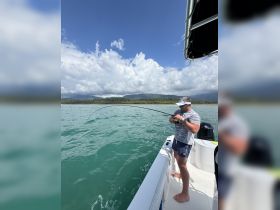 A fisherman enjoys a day on the water, casting his line from the side of a boat against a backdrop of lush mountains and clear skies — perfect for adventure seekers and fishing enthusiasts.