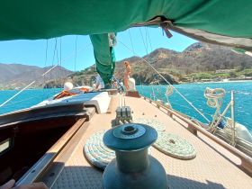 A scenic view from the deck of a sailboat, showcasing the vibrant blue ocean and a guest enjoying the sailing experience against a backdrop of lush hills.