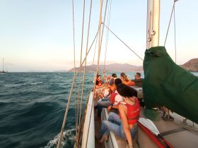 A lively group enjoys a sailing experience at sunset, wearing life jackets and taking in the beautiful coastal scenery.