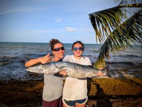 Two women proudly display their catch of the day, showcasing a large fish on the beach — a memorable moment from a successful fishing trip.