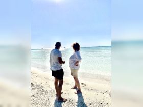A couple enjoys a serene moment on the beach, gazing at the tranquil waters and distant boats — a perfect snapshot of relaxation and connection with nature.