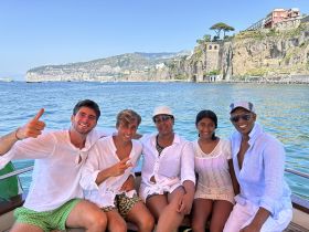 A cheerful group of friends poses for a photo on a boat, enjoying a sunny day on the water with stunning coastal views in the background.